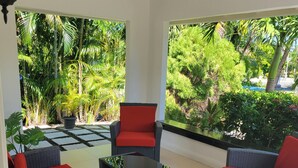 A covered patio area with wicker furniture and red cushions, overlooking a lush garden with various plants and trees. - Island Treasure Villa (St. Ann, Ocho Rios)