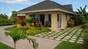 A single-story house with a tiled roof, a front porch, and a well-maintained garden with various plants and a palm tree. - Island Treasure Villa (St. Ann, Ocho Rios)