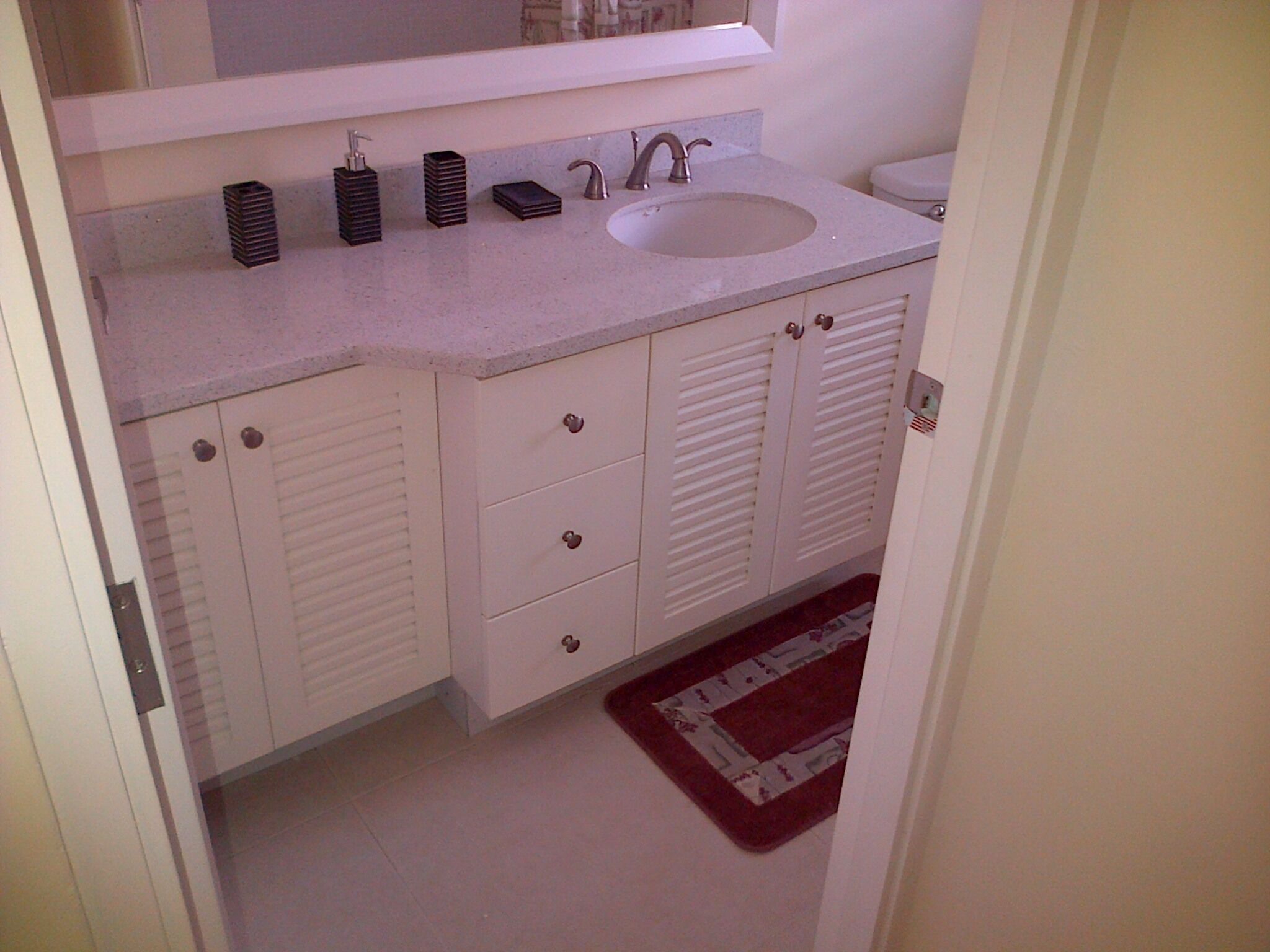 A bathroom with a white sink, two faucets, a toilet, and a small mirror above the sink. There are three drawers and two cabinets below the sink.