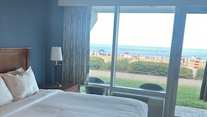 Een hotelkamer met een groot bed, een nachtkastje, een lamp en uitzicht op het strand door een groot raam.