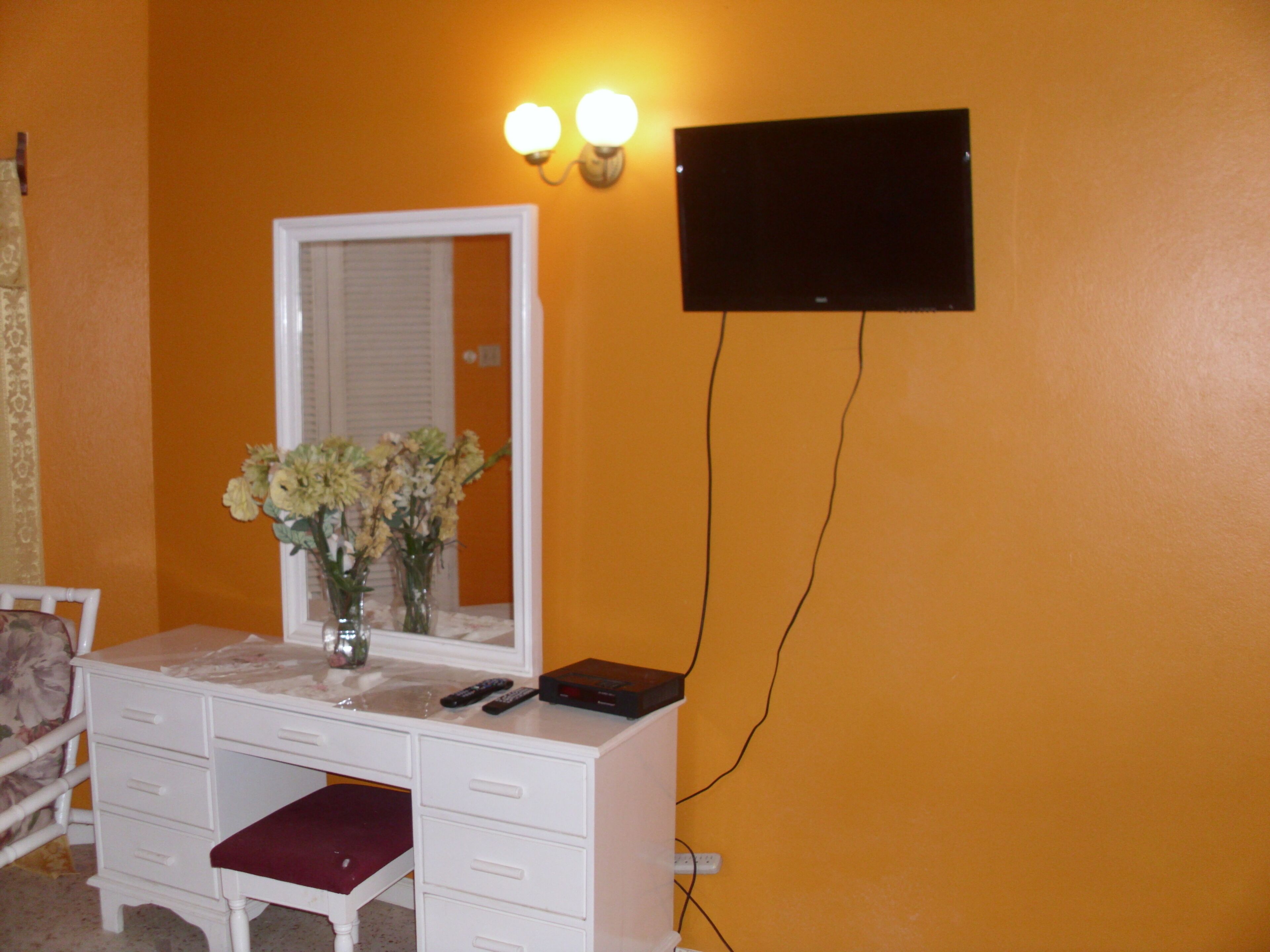 A room with a white vanity, a mirror, and a vase of flowers. There is a black flat-screen TV mounted on the wall.