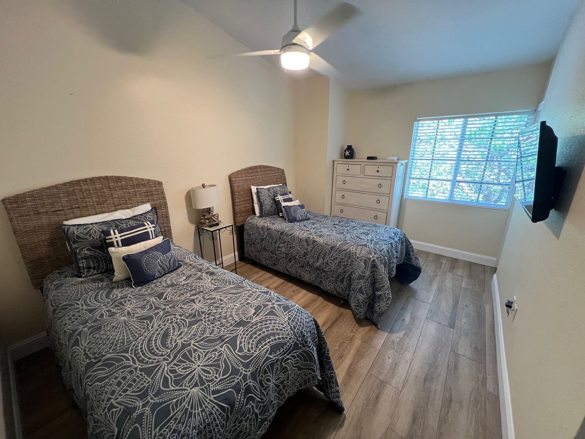 Une chambre avec deux lits, un ventilateur de plafond, une commode, une fenêtre avec des stores et un téléviseur fixé au mur.