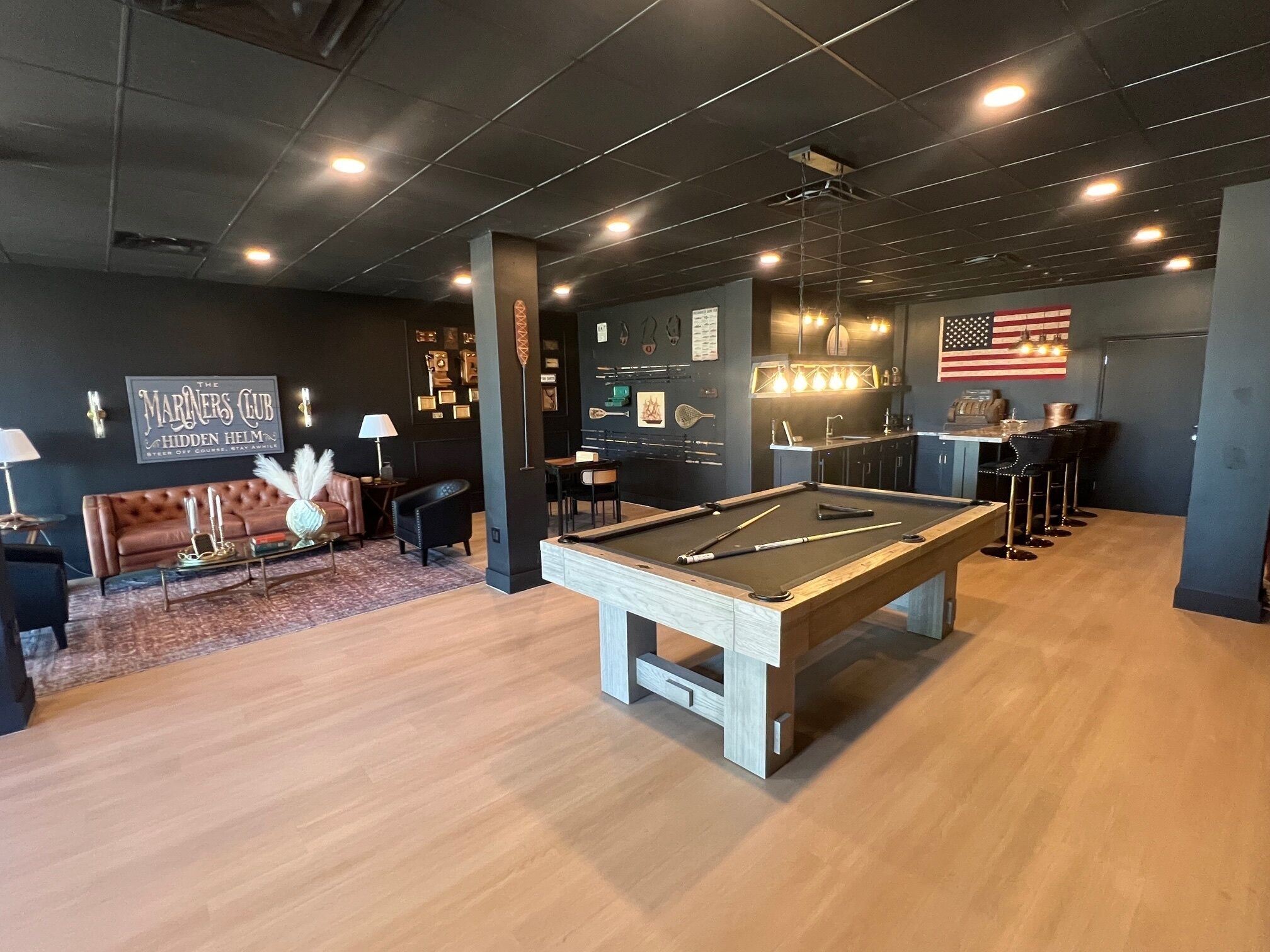 Une table de billard dans une pièce aux murs et au plafond sombres, un coin bar avec des tabourets et une enseigne indiquant : « The Mariner's Club Hidden Helm ».