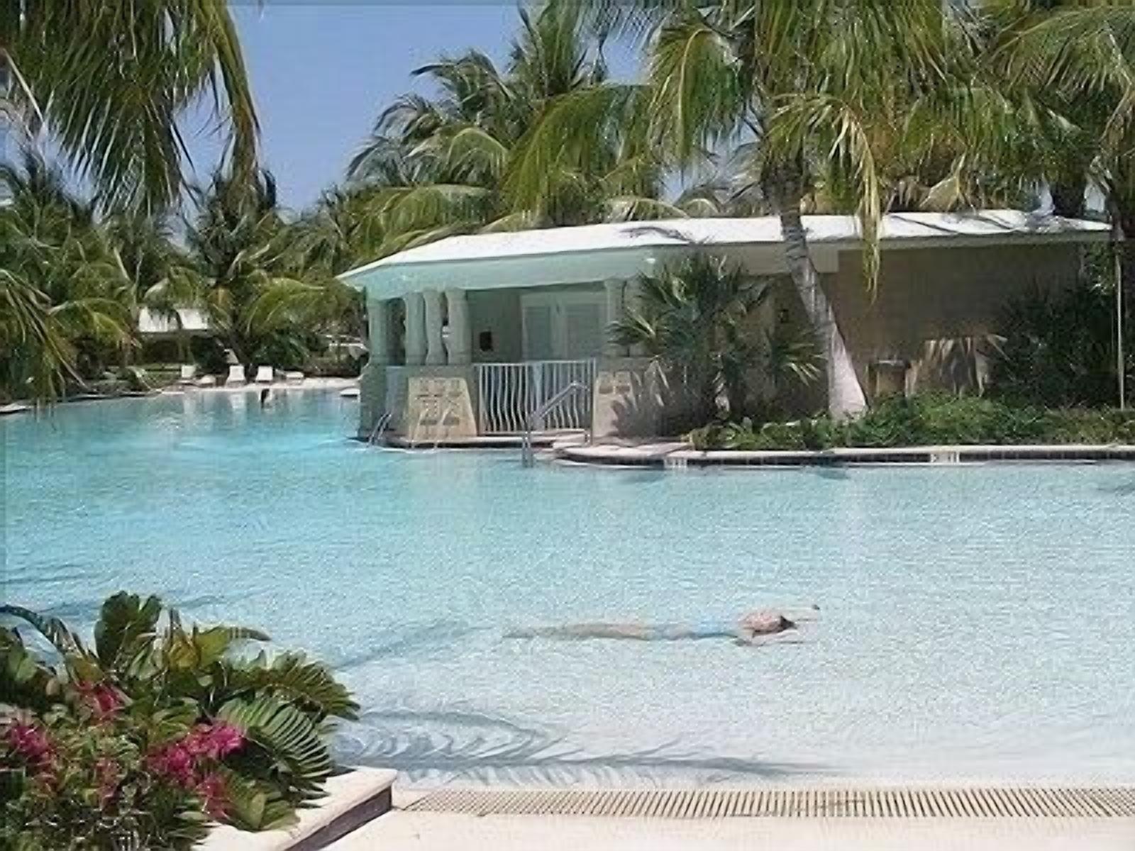 A clear swimming pool with a lifeguard tower, surrounded by palm trees and a building with columns.