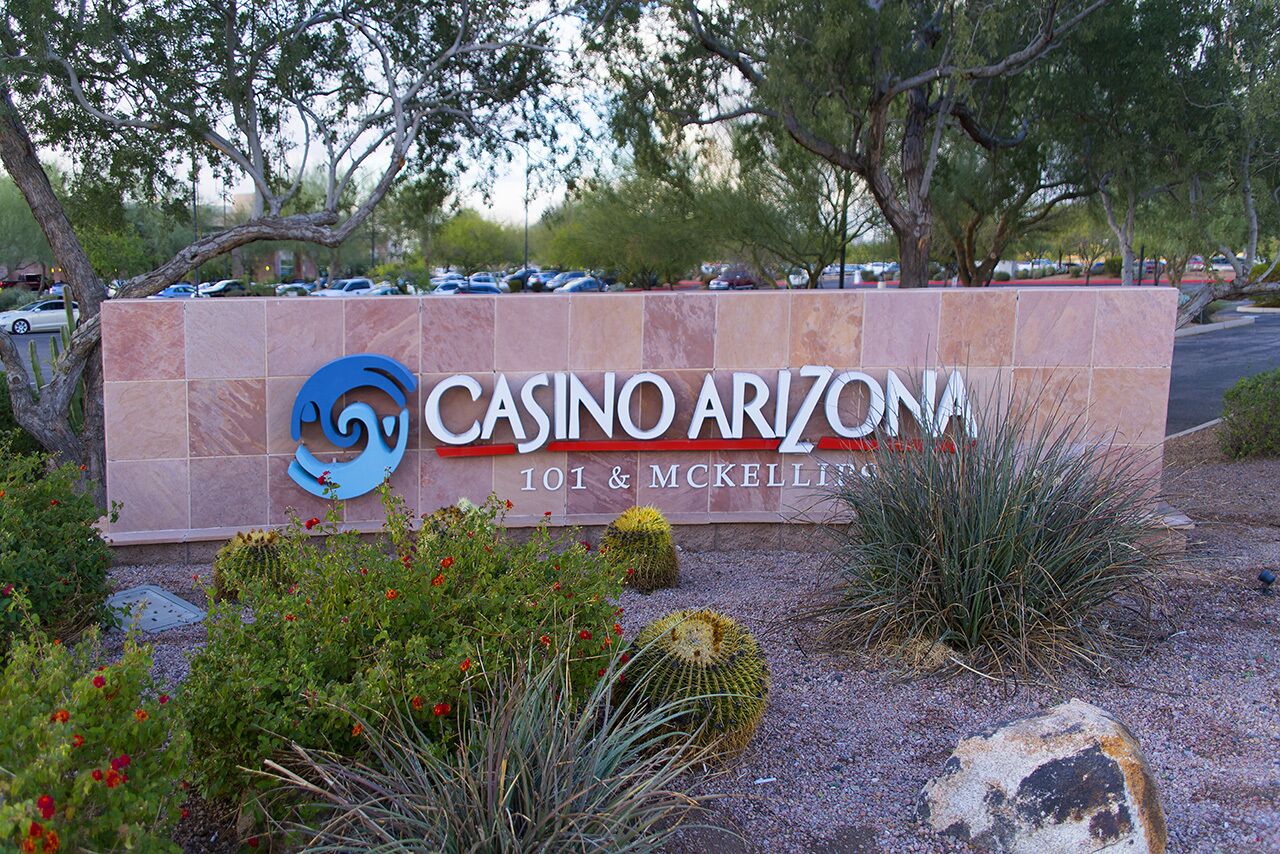 A sign for 'Casino Arizona 101 & McKellin' with a logo, surrounded by desert plants and trees.