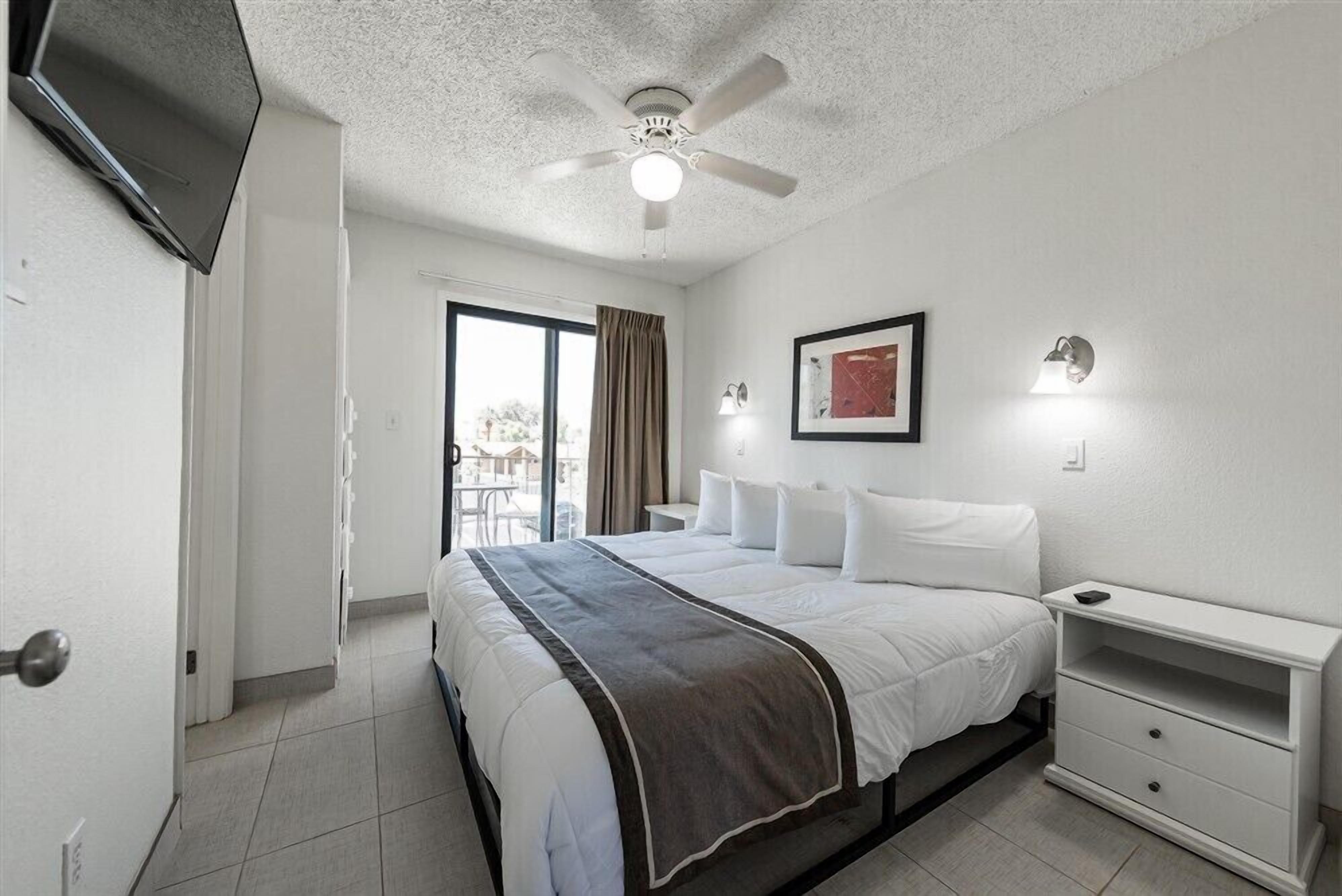 Een hotelkamer met een bed, nachtkastje, plafondventilator, televisie en raam met gordijnen.
