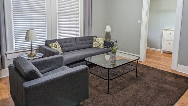 Ein Wohnzimmer mit einem grauen Modulsofa, einem Glastisch und einem dunkelbraunen Teppich.