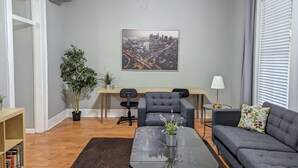Ein modernes Wohnzimmer mit einer Couch, einem Glastisch, einem Schreibtisch und einem Sessel. An der Wand hängt ein Stadtbild, es gibt einen Deckenventilator und ein Fenster mit Jalousien.