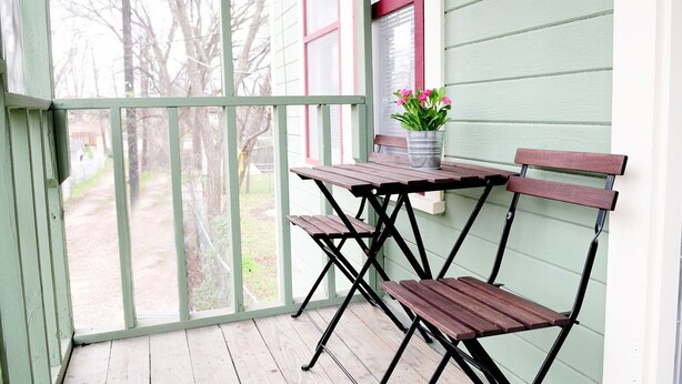 Une véranda abritée avec une table en bois et deux chaises, un pot de fleurs et une vue sur une cour avec des arbres.