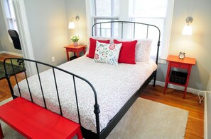 Ein ordentlich eingerichtetes Schlafzimmer mit einem Bett, Nachttischen, einer Bank und einem Fenster mit Jalousien.