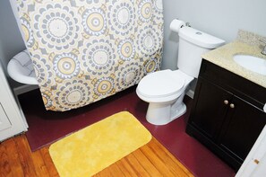Ein Badezimmer mit einem gemusterten Duschvorhang, einer weißen Toilette, einem Waschbecken mit einer Granit-Arbeitsplatte und einem Holzboden.