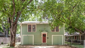 Une maison à deux étages avec une porte rouge et un revêtement extérieur vert, entourée d’arbres et d’une cour clôturée.