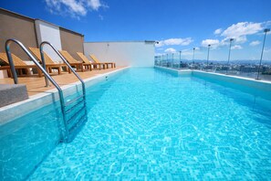 A rooftop swimming pool with clear blue water, wooden lounge chairs, and a city view.