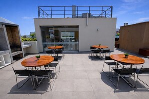 A rooftop patio with four wooden tables and chairs, a glass door entrance, and a modern building in the background.