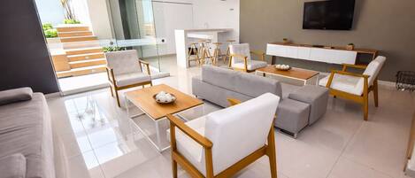 A modern living room with a sofa, chairs, a coffee table, a TV, and a kitchen area.