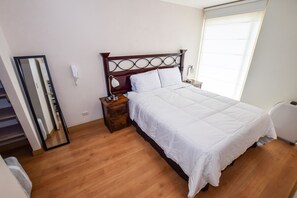 A bedroom with a wooden floor, a bed with white bedding, a nightstand with a lamp, a mirror, and a window with blinds.