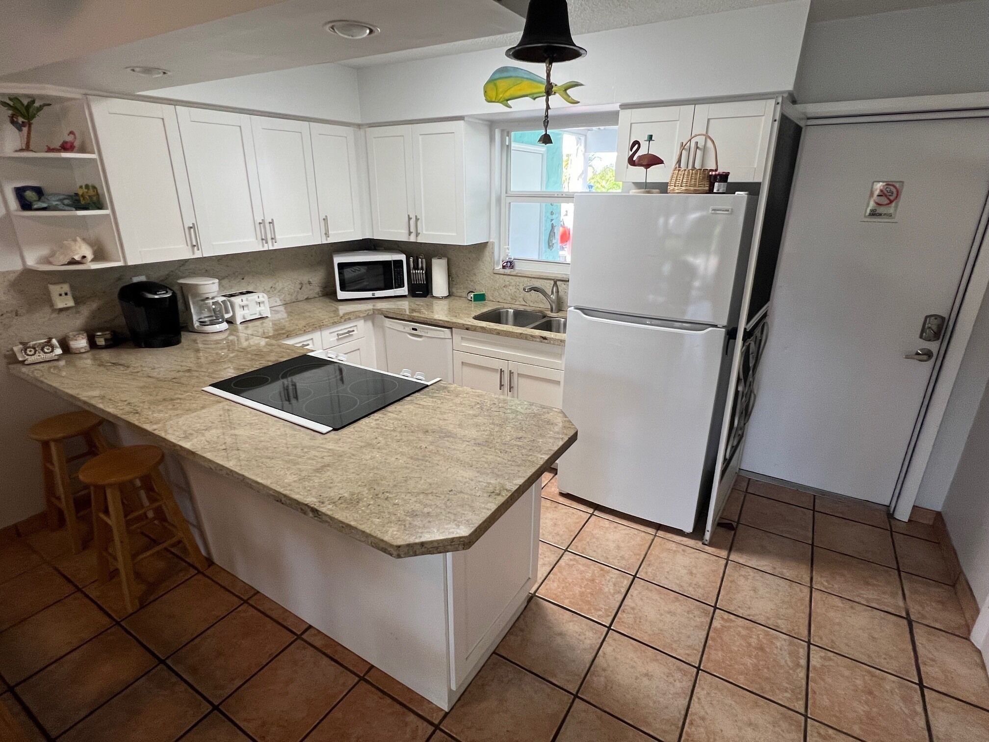 A kitchen with white cabinets, a microwave, a refrigerator, a sink, and a window with a hanging decoration.