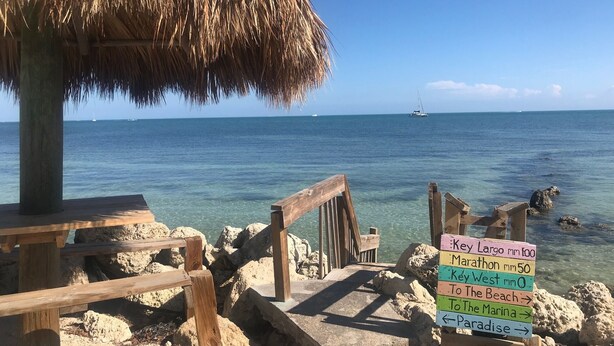 Une jetée en bois surmontée d’un toit de chaume, qui mène à une plage rocheuse aux eaux d’un bleu limpide. Des panneaux indicateurs signalent les lieux à proximité.