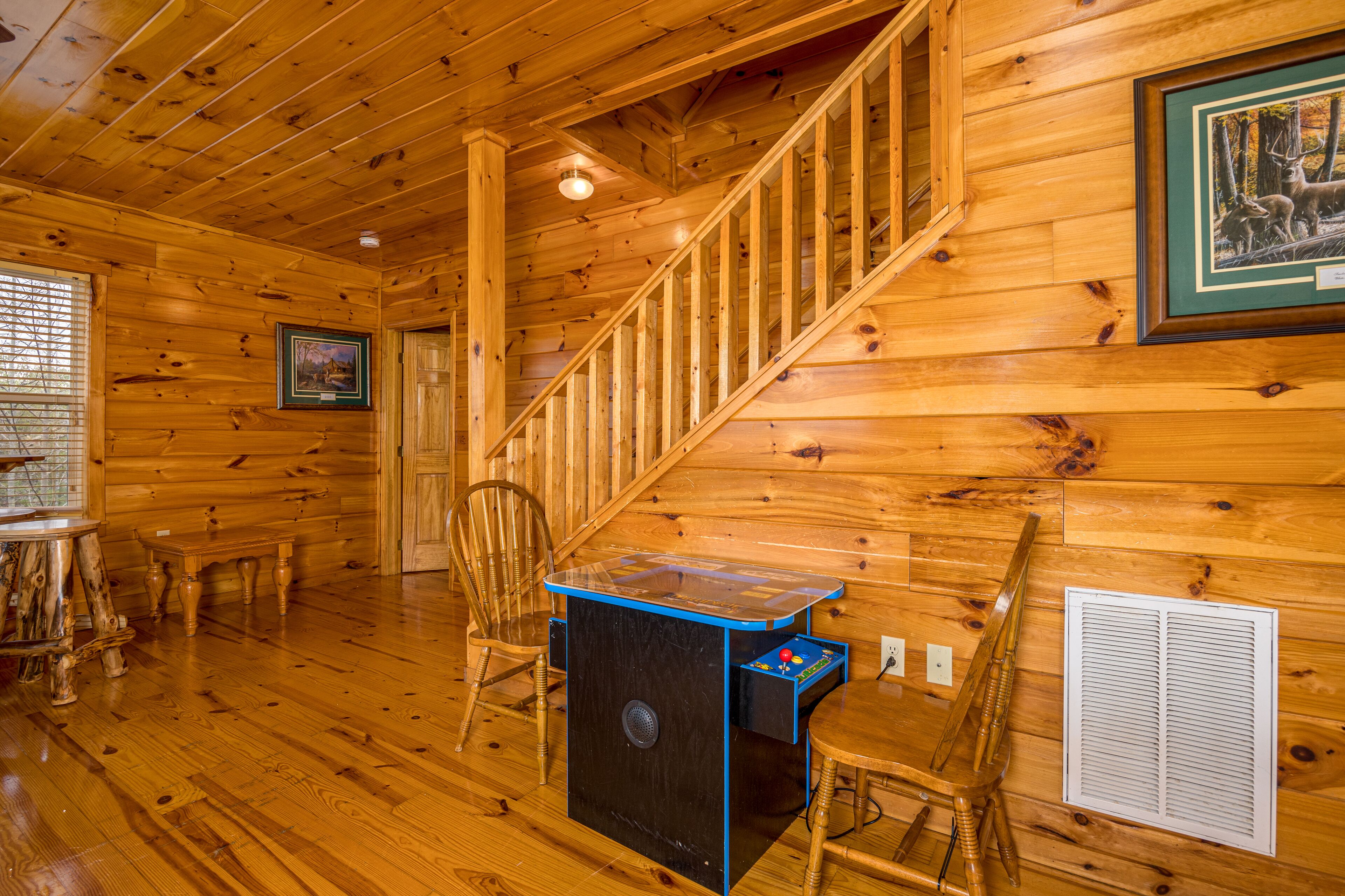Interior de una cabaña de madera con una escalera, una pequeña mesa con superficie azul, una silla y un cuadro en la pared.