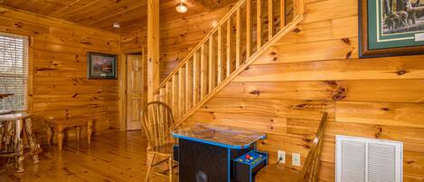 Interior de una cabaña de madera con una escalera, una pequeña mesa con superficie azul, una silla y un cuadro en la pared.
