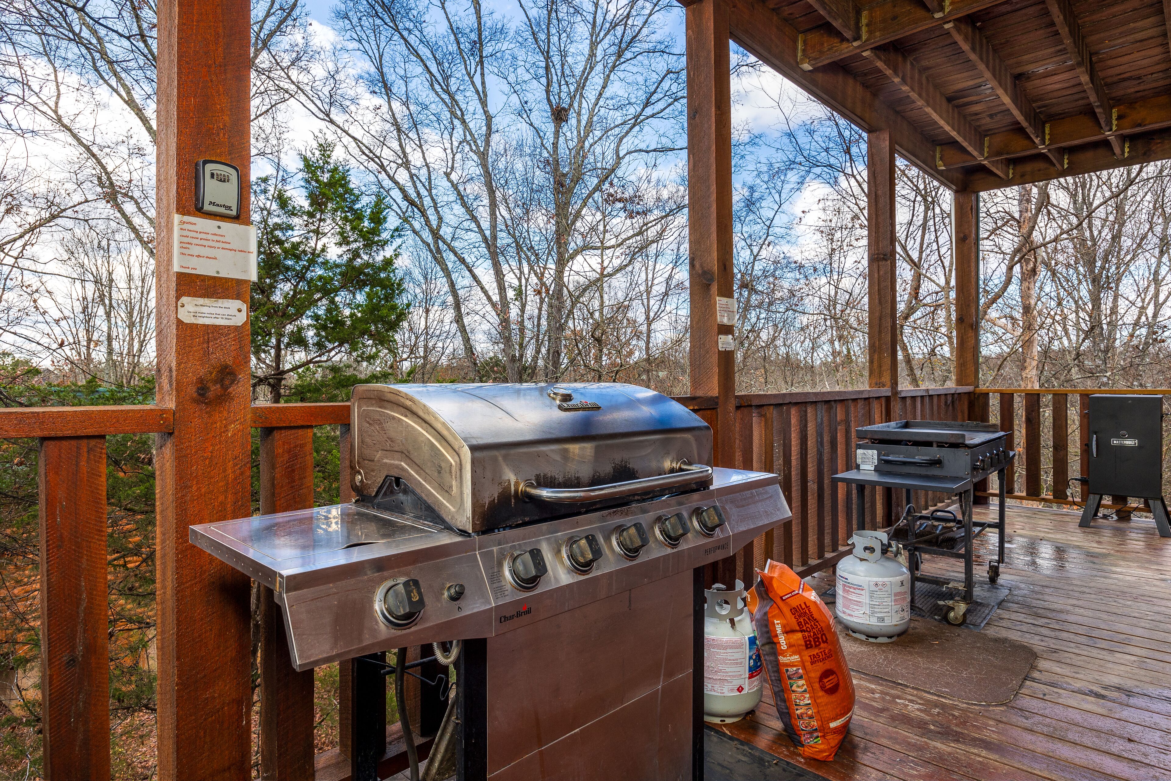 A barbecue grill with a digital thermometer, a gas tank, and a grill cart on a wooden deck with a view of trees.