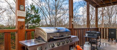 A barbecue grill with a digital thermometer, a gas tank, and a grill cart on a wooden deck with a view of trees.