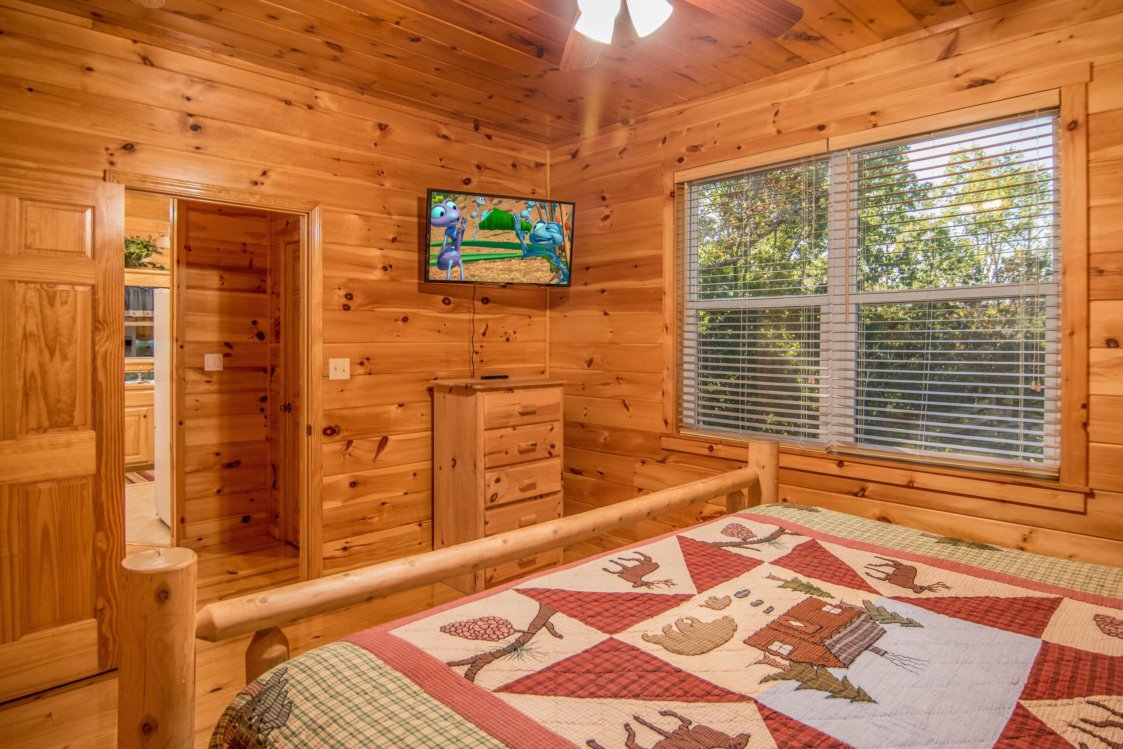 A wooden cabin interior with a bed, a television, a window with blinds, and a door leading to another room.