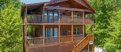 A two-story log cabin with a balcony on the upper floor, surrounded by lush greenery.