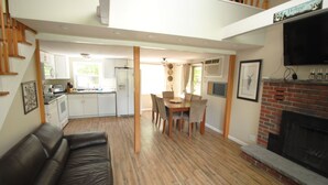 Uma sala de estar ampla com piso de madeira, um sofá de couro preto, uma mesa de jantar com cadeiras, uma cozinha ao fundo e uma lareira.