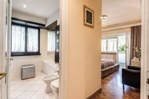 A bathroom with a toilet, bidet, and bathtub, a bedroom with a bed and chair, and a hallway with a door leading to the bathroom.
