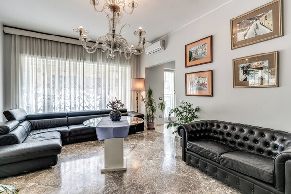 A modern living room with a black leather sofa, a round coffee table, a chandelier, and framed pictures on the wall.