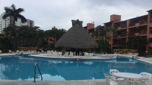 Une piscine entourée de chaises longues et surmontée d’une structure à toit de chaume, le tout bordé de palmiers et de bâtiments.