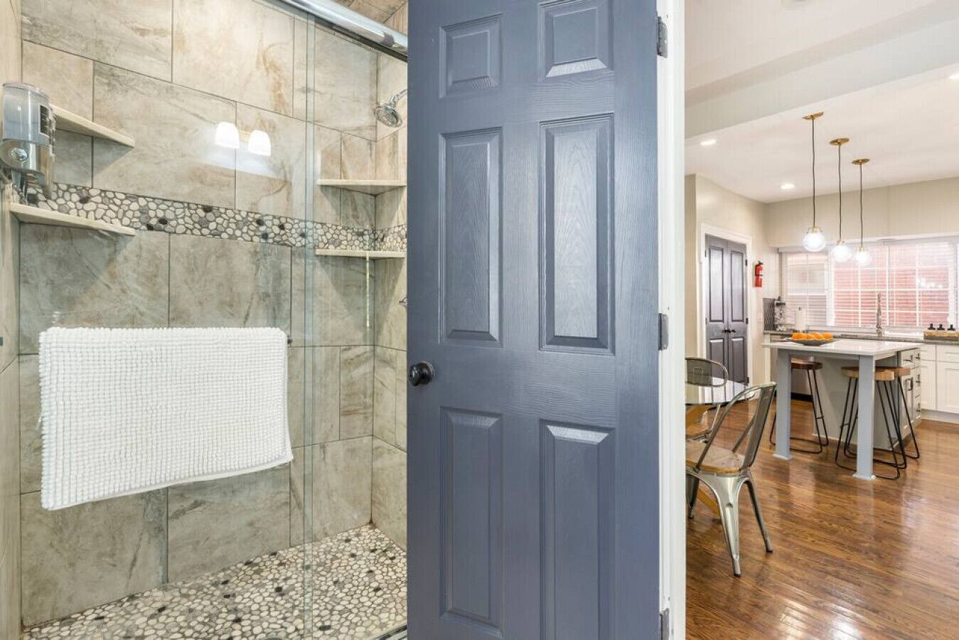 A modern bathroom with a shower, a towel rack, and a door leading to a kitchen area with a dining table and bar stools.