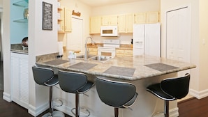 Una cocina moderna con encimeras de granito, cuatro taburetes de bar, un microondas, un refrigerador y armarios de madera.