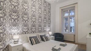 Una camera da letto con carta da parati decorata, un letto con cuscini, un comodino con una lampada e una finestra che si affaccia sulla città.