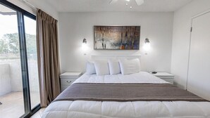 Un dormitorio con una cama grande, ventilador de techo, un cuadro sobre la cama y vista a través de un ventanal corredero.