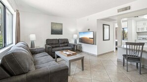 A modern living room with a grey sofa, a coffee table, a TV, and a kitchen area visible through an open doorway. - 133 Fully Furnished, amenities galore! (Scottsdale)