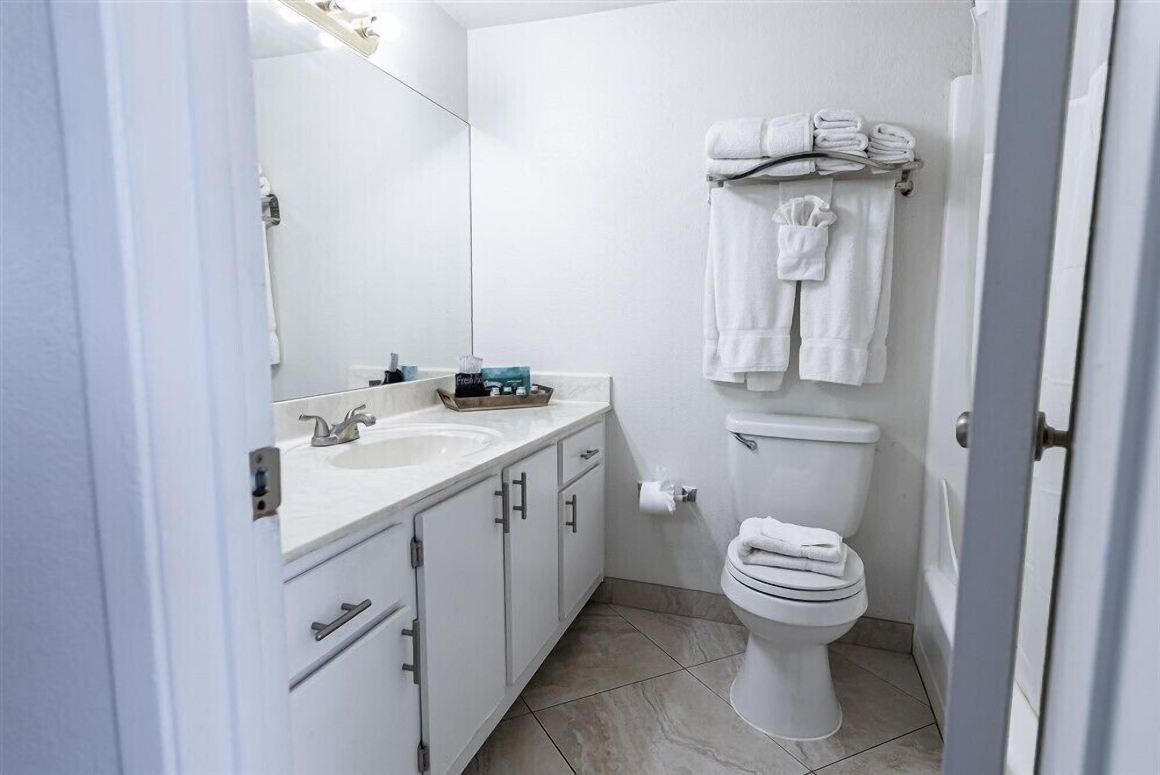 A modern bathroom with a white sink, mirror, and toilet. There are towels hanging on a rack and a small shelf with toiletries.