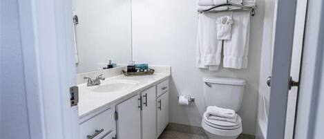 A modern bathroom with a white sink, mirror, and toilet. There are towels hanging on a rack and a small shelf with toiletries.