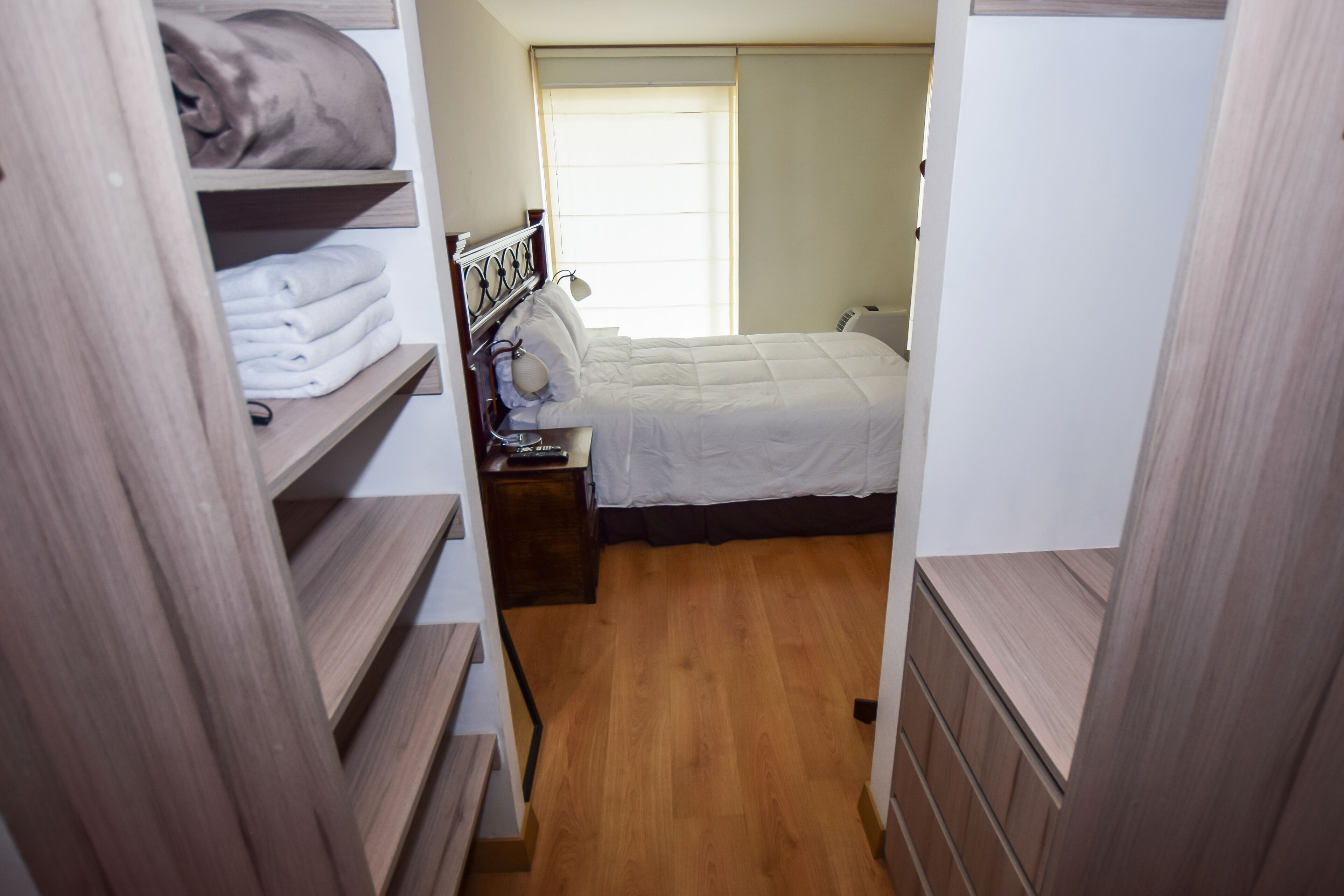 Une chambre à coucher avec un lit, une table de chevet et une fenêtre. Il y a un placard avec des étagères et une porte.