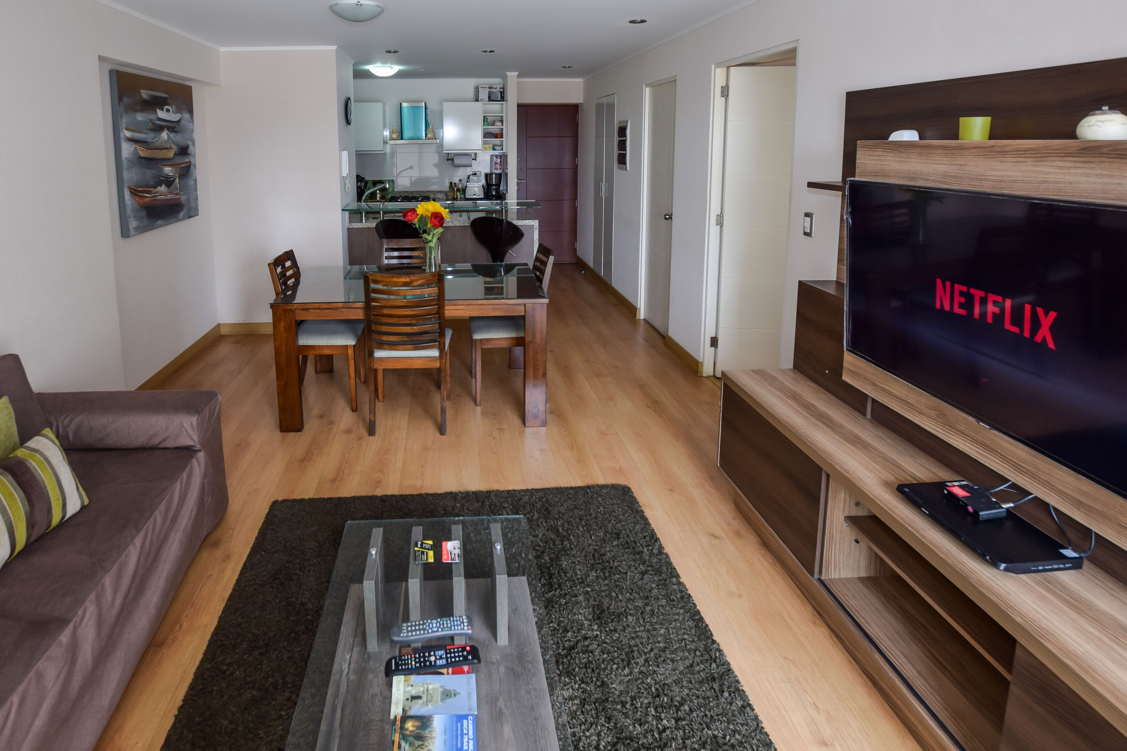 Ein modernes Wohnzimmer mit Flachbildfernseher, Holzmöbeln und einem Glastisch mit Fernbedienungen.
