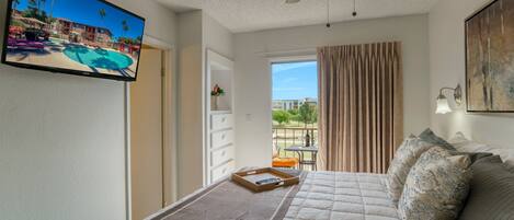 A bedroom with a bed, a ceiling fan, a TV, and a view of a pool and building through a window.