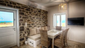 Ein Zimmer mit Steinwand, einer weißen Tür, einem weißen Tisch mit Korbstühlen, einer Bank mit Kissen und einem Fernseher an der Wand.
