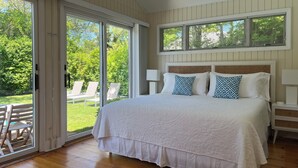 A bedroom with a large bed, a wooden floor, and a sliding glass door leading to an outdoor area with chairs. - Stylish East Hampton Village Retreat- Heated Pool and Sauna, Steps to Village (East Hampton)