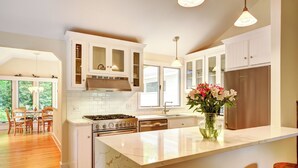 A modern kitchen with white cabinetry, a central island, and a dining area with wooden chairs. - Stylish East Hampton Village Retreat- Heated Pool and Sauna, Steps to Village (East Hampton)