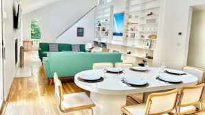 Ein moderner Essbereich mit einem runden Tisch, gedeckt für sechs Personen, umgeben von sechs Stühlen. Ein grünes Sofa befindet sich im Hintergrund, und eine Treppe führt auf eine höhere Ebene.