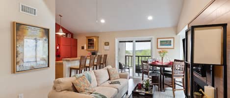 A spacious living area with a sofa, coffee table, and dining table. There is a kitchen area with cabinets and a dining set. A ceiling fan and a large window are visible.