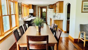 A spacious dining area with a long wooden table and chairs, flanked by wooden cabinets and large windows.