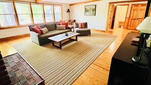 A spacious living room with a large sofa, a coffee table, and a rug. There are large windows providing natural light and a view of the outdoors.