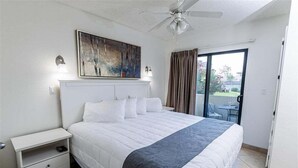 Ein Schlafzimmer mit Bett, Nachttisch, Deckenventilator, einem Gemälde und einer Glastür zum Außenbereich.
